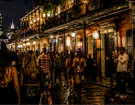 A nighttime street party with people in costumes watching a brass band play, while two women wear witch hats in the foreground.