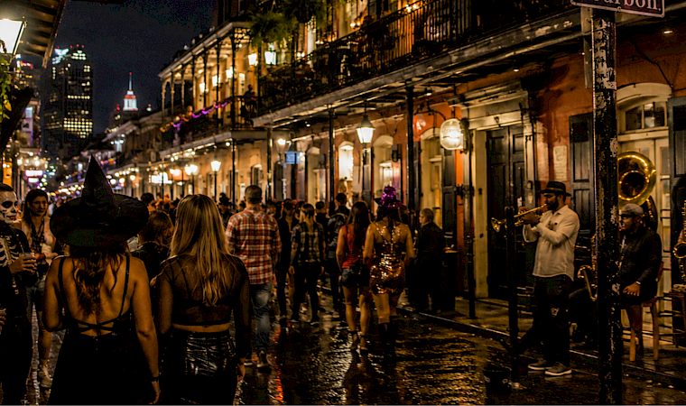 A nighttime street party with people in costumes watching a brass band play, while two women wear witch hats in the foreground.