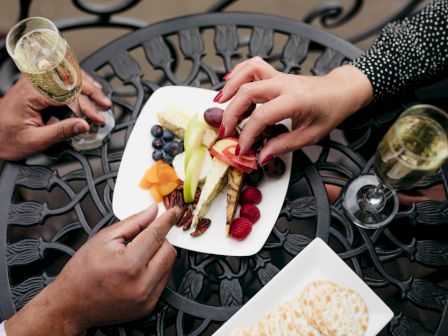 A cheese-and-fruit platter is being shared at a table, with sparkling drinks in glasses and hands handing pieces over.