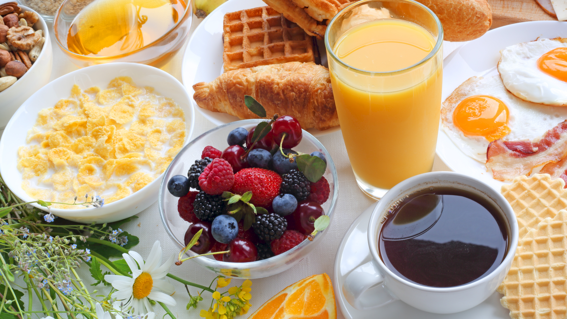 A hearty breakfast spread with waffles, croissants, eggs, yogurt, fruit bowl, juice, and coffee.