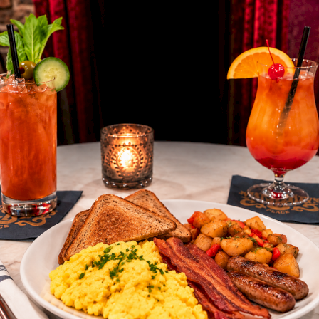A brunch platter with scrambled eggs, sausage, bacon, roasted potatoes, plus two colorful cocktails on the table.