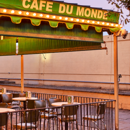 &ldquo;Cafe du Monde&rdquo; outdoor patio with a green awning, glowing bulbs, and metal chairs waiting for coffee and beignets.