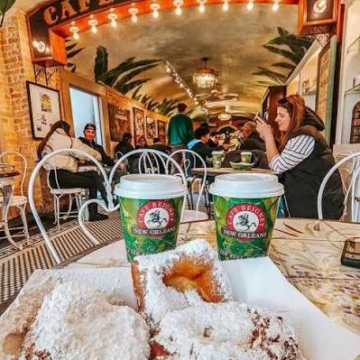 Beignet caf&eacute;: powdered-sugar beignets on a table with two caf&eacute; cups and a &ldquo;Cafe Beignet&rdquo; sign in view.