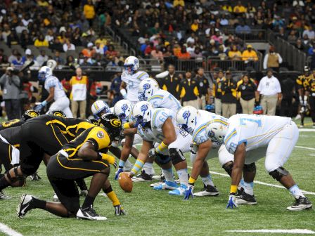 An American football game is underway with players in formation at the line of scrimmage, preparing for the snap.