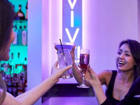 Two women clink glasses with drinks at a neon-lit bar, laughing and celebrating together.