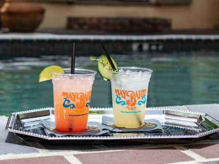 Two iced drinks from &ldquo;MayBaily&rsquo;s Place&rdquo; sit poolside on a tray&mdash;one orange and one pale yellow with lime and herbs.