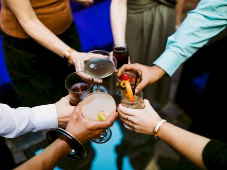 Friends clink glasses in a celebratory toast with cocktail drinks and colorful garnishes.