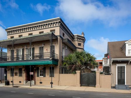 A sunny street scene featuring a historic, multi-story building with arched balconies, wrought-iron railings, and palm-like plants.