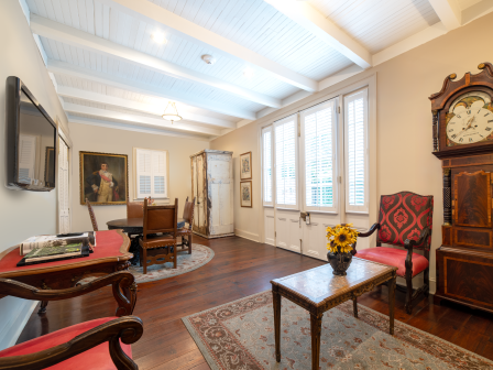 An elegant parlor with tall wooden grandfather clock, framed paintings, leather chairs, warm lamps, a TV, and large shuttered windows.